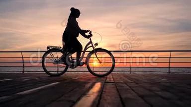 美妙的日出或日落在海洋之上。 年轻时尚女孩骑着老式自行车在木堤上骑车剪影